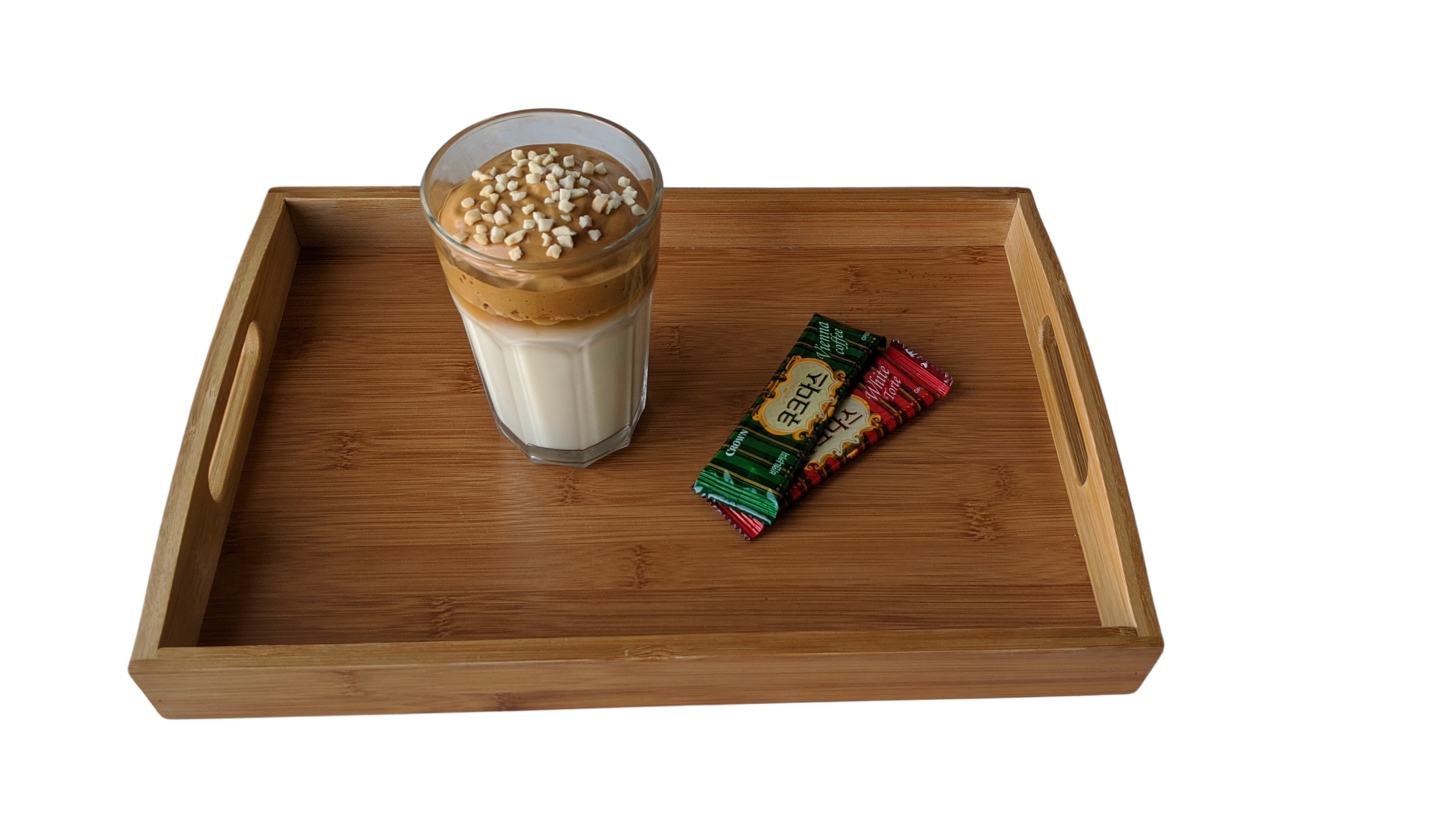 Glas Dalgona-Kaffee mit aufgeschlagener Kaffeecreme über Milch und Nussstückchen, daneben zwei koreanische Instantkaffee-Sticks auf einem Bambustablett