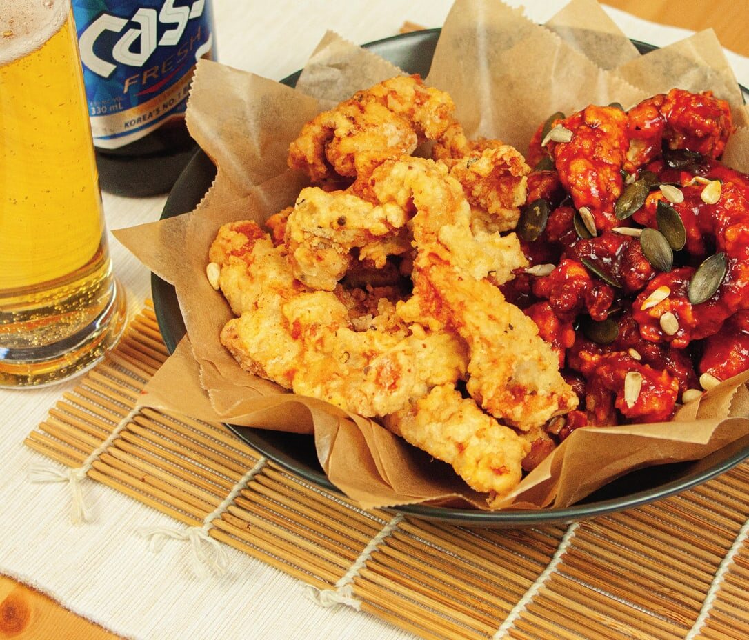 Dak-Gangjeong auf Backpapier in einer Schale, links pur frittiert, rechts in roter Soße mit Kürbis- und Sonnenblumenkernen, daneben ein Glas Bier