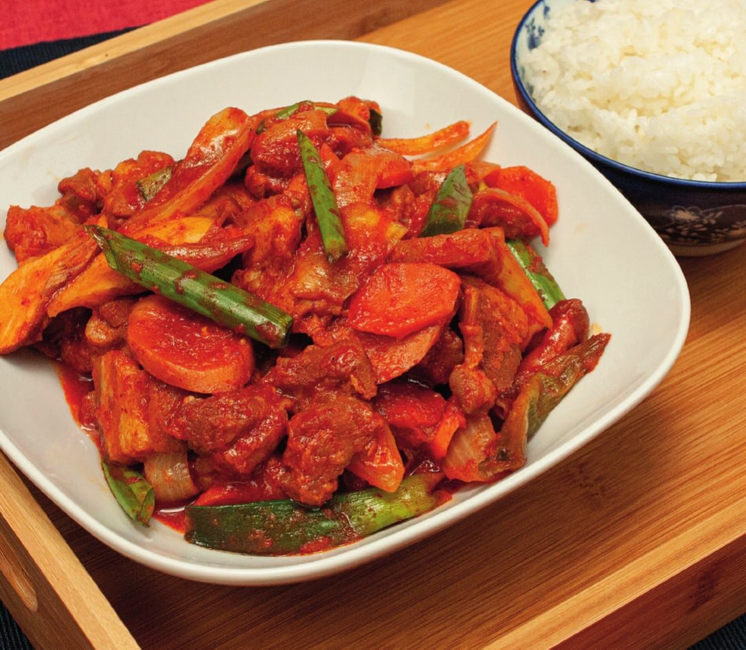 Jeyuk-Bokkeum mit Schweinefleisch, Karotten, Pilzen und Frühlingszwiebeln auf einem Teller, daneben eine Schale Reis