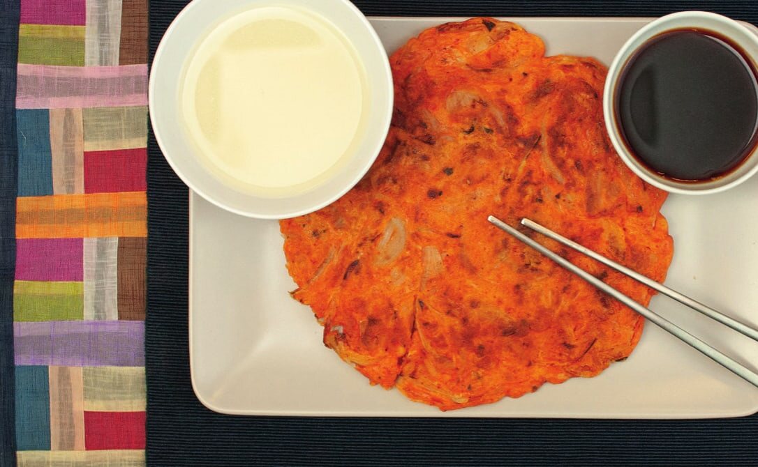 Kimchi-Jeon auf einem eckigen Teller mit Stäbchen, daneben eine Schale Makgeolli und eine Schale Sojasoße