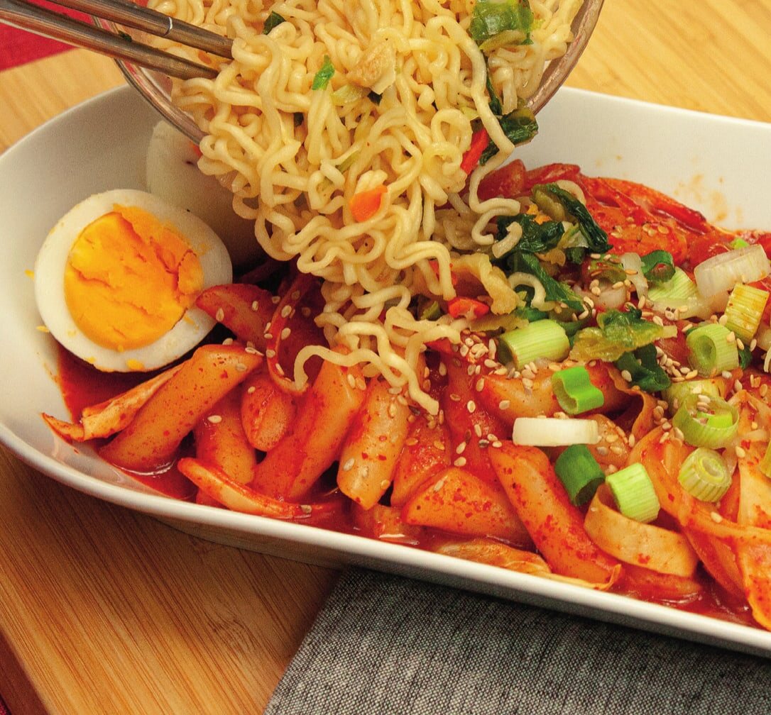 Instant-Ramen werden mit Stäbchen auf Tteokbokki in roter Soße gegeben, daneben ein halbes Ei, Frühlingszwiebeln und Sesam