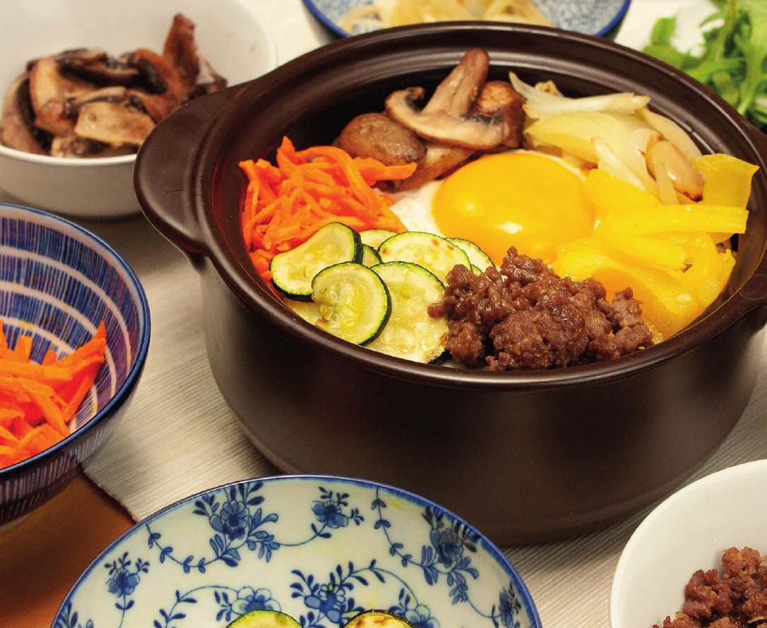 Bibimbap im Tontopf mit Spiegelei, Karotten, Zucchini, Hackfleisch, Pilzen und Paprika, ringsum Schälchen mit Zutaten
