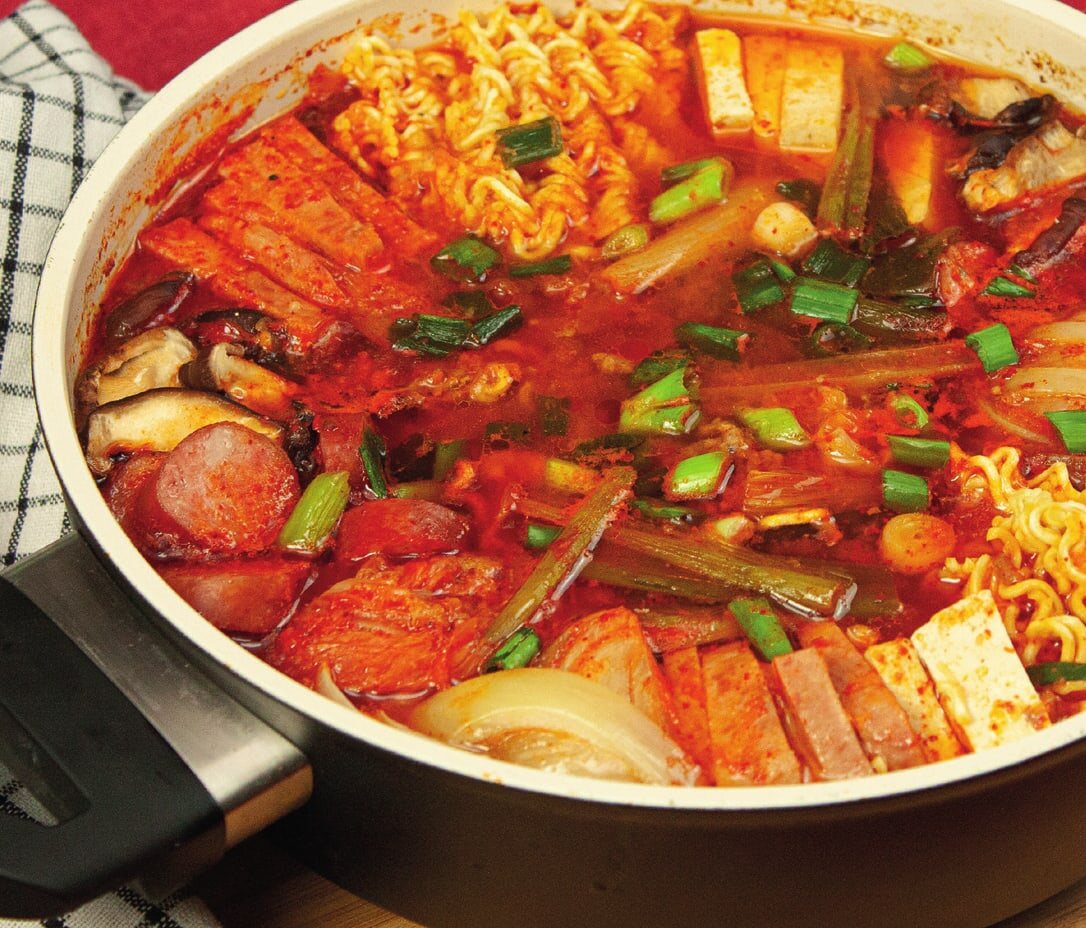 Budae-Jjigae im Topf mit Ramen-Nudeln, Würstchen, Frühstücksfleisch, Tofu, Pilzen und Frühlingszwiebeln in roter Brühe