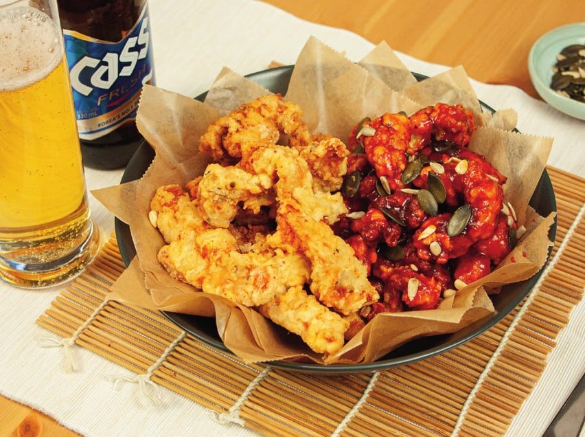 Fertiges Dak-Gangjeong, eine Hälfte pur, die andere in roter Soße mit Kernen, daneben Bier