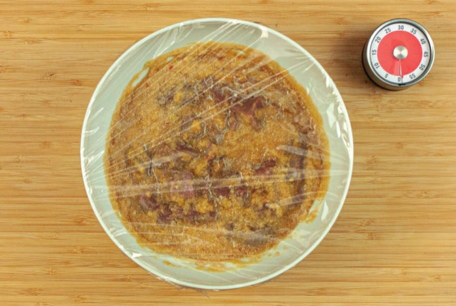 Mit Folie abgedeckte Schüssel mit Fleisch in Marinade, daneben ein Küchenwecker