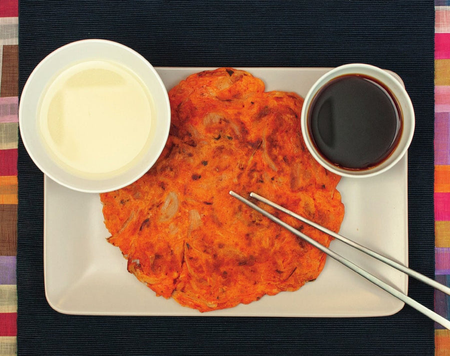 Fertiger Kimchi-Jeon auf einem eckigen Teller mit Stäbchen, daneben Makgeolli und Sojasoße