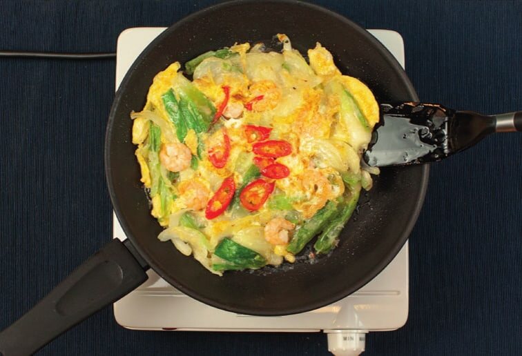 Pajeon in der Pfanne mit Garnelen, Ei und Chiliringen, daneben ein Pfannenwender