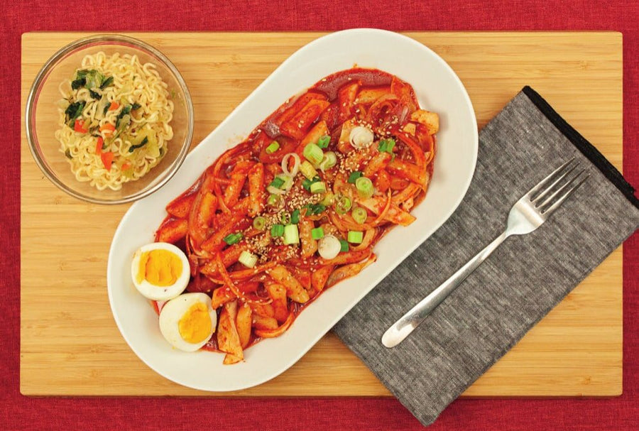 Tteokbokki mit Eihälften, Frühlingszwiebeln und Sesam auf einer ovalen Platte, daneben eine Schale Ramen-Nudeln